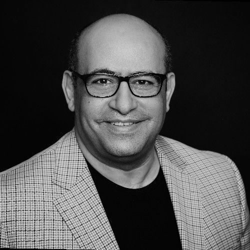 Smiling man with glasses, wearing a patterned blazer against a dark background.